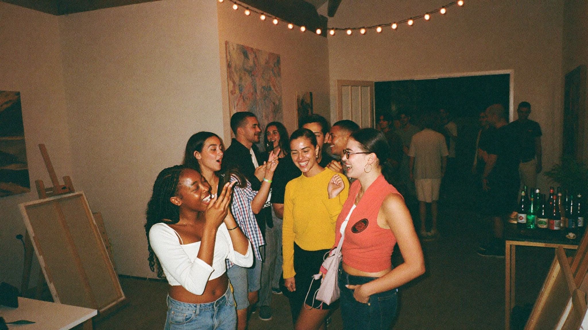 A packed creator-hosted pop-up event inside an art studio, crowd talking and filming, drinks table in the corner